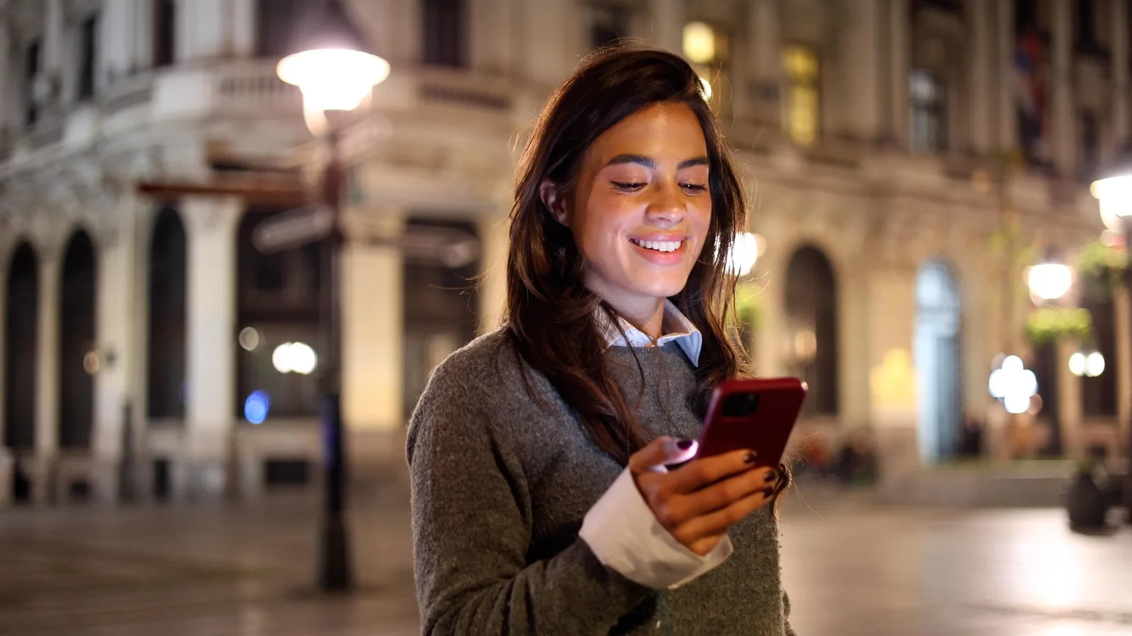 A young woman smiling at her phone on a lit city street at night